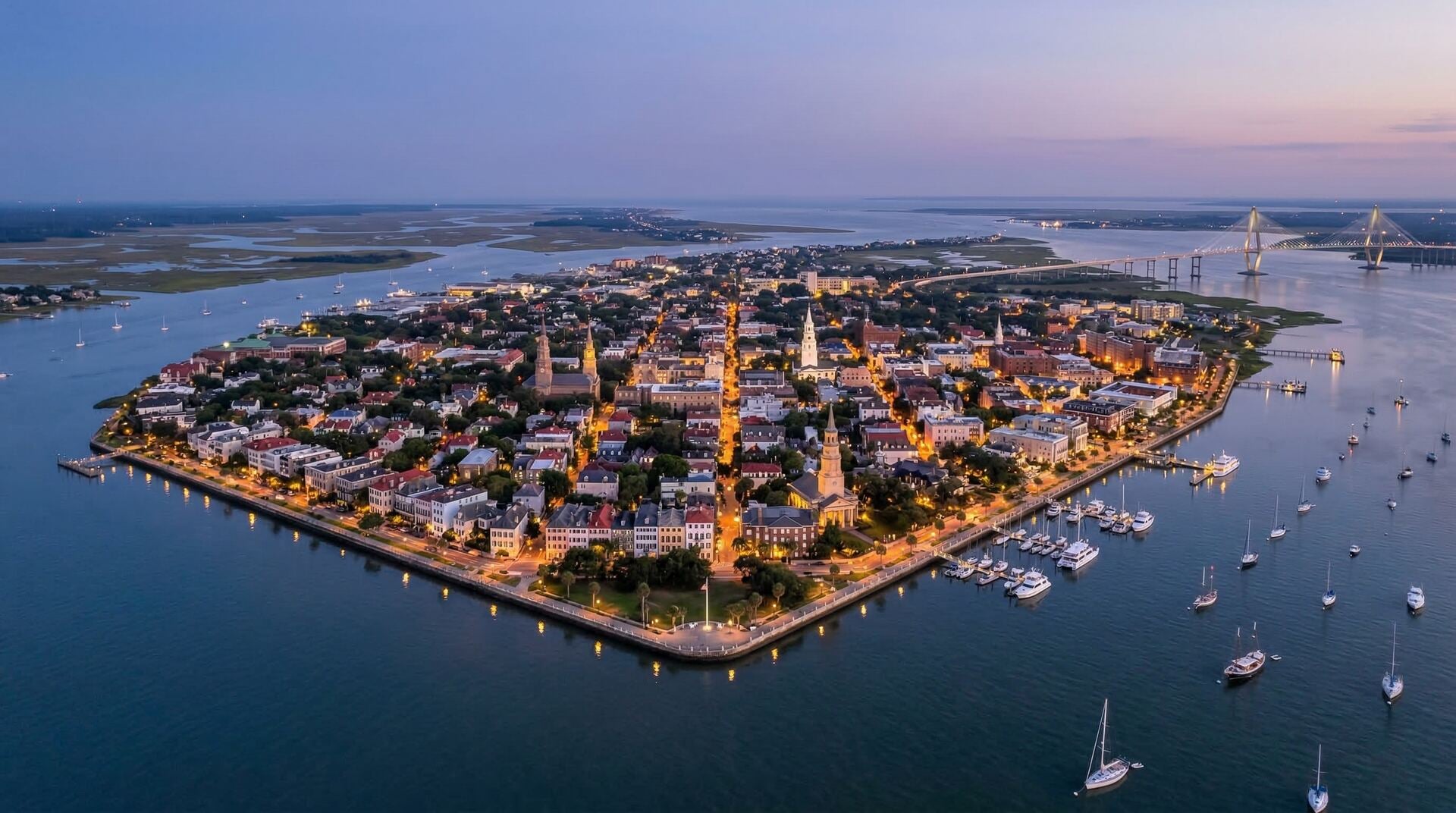 Aerial view of the Charleston peninsula at twilight with church steeples, lit streets, and harbor